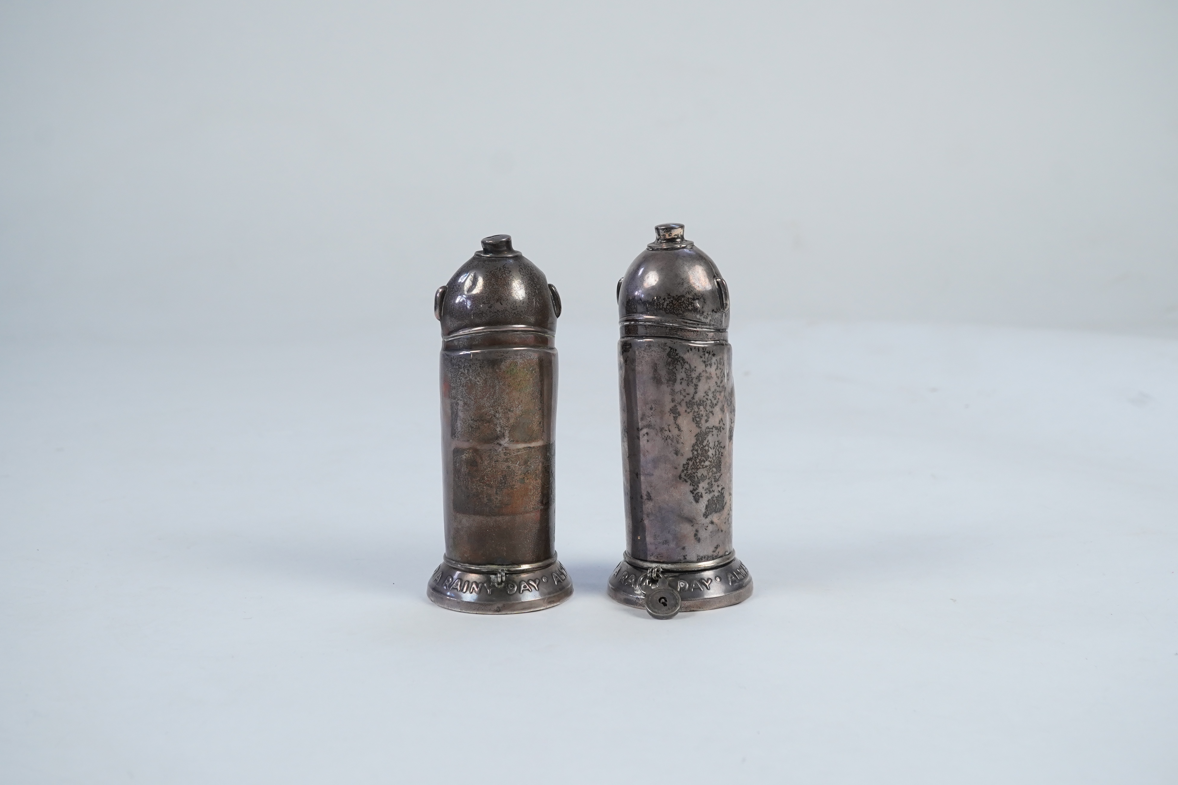 A matched pair of George V novelty silver money boxes, each cylindrical box embossed as a banker with an umbrella, the circular base inscribed 'Always be prepared for a rainy day'